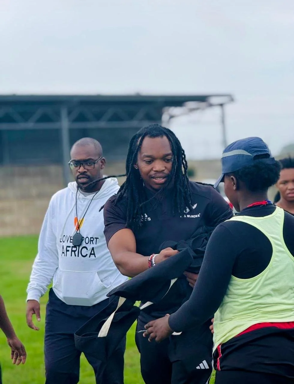 A man wearing a black sports shirt shaking hands with a woman in a yellow vest during an outdoor sports event, with another man observing in the background.