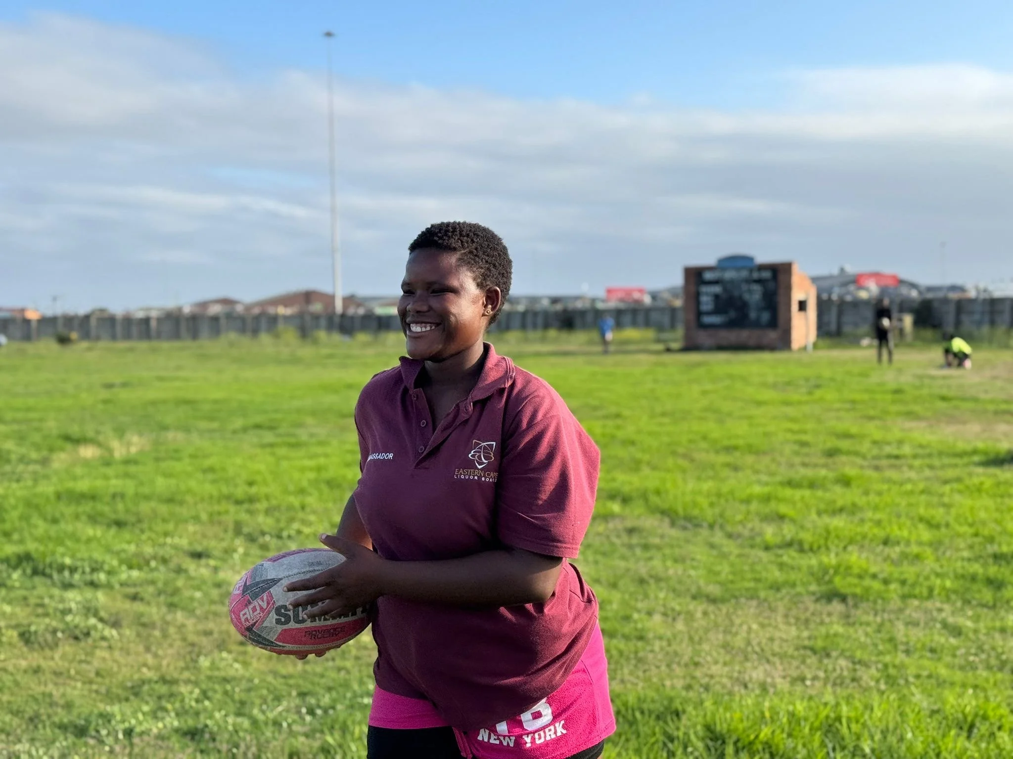A woman smiling and holding a rugby ball on a grassy field with a fence, structures, and a digital scoreboard in the background on a cloudy day.
