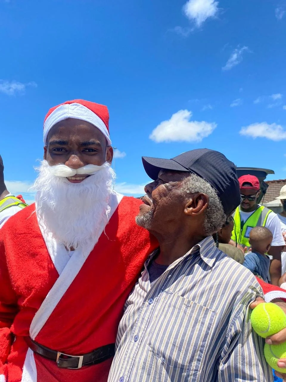 Three people standing outdoors on a cloudy day, smiling, with their arms around each other. The woman on the left and the man on the right are wearing green T-shirts with a logo that says 'Love for Africa.' The man in the middle is wearing a black shirt, a Santa hat, and shorts with red, white, and blue colors.