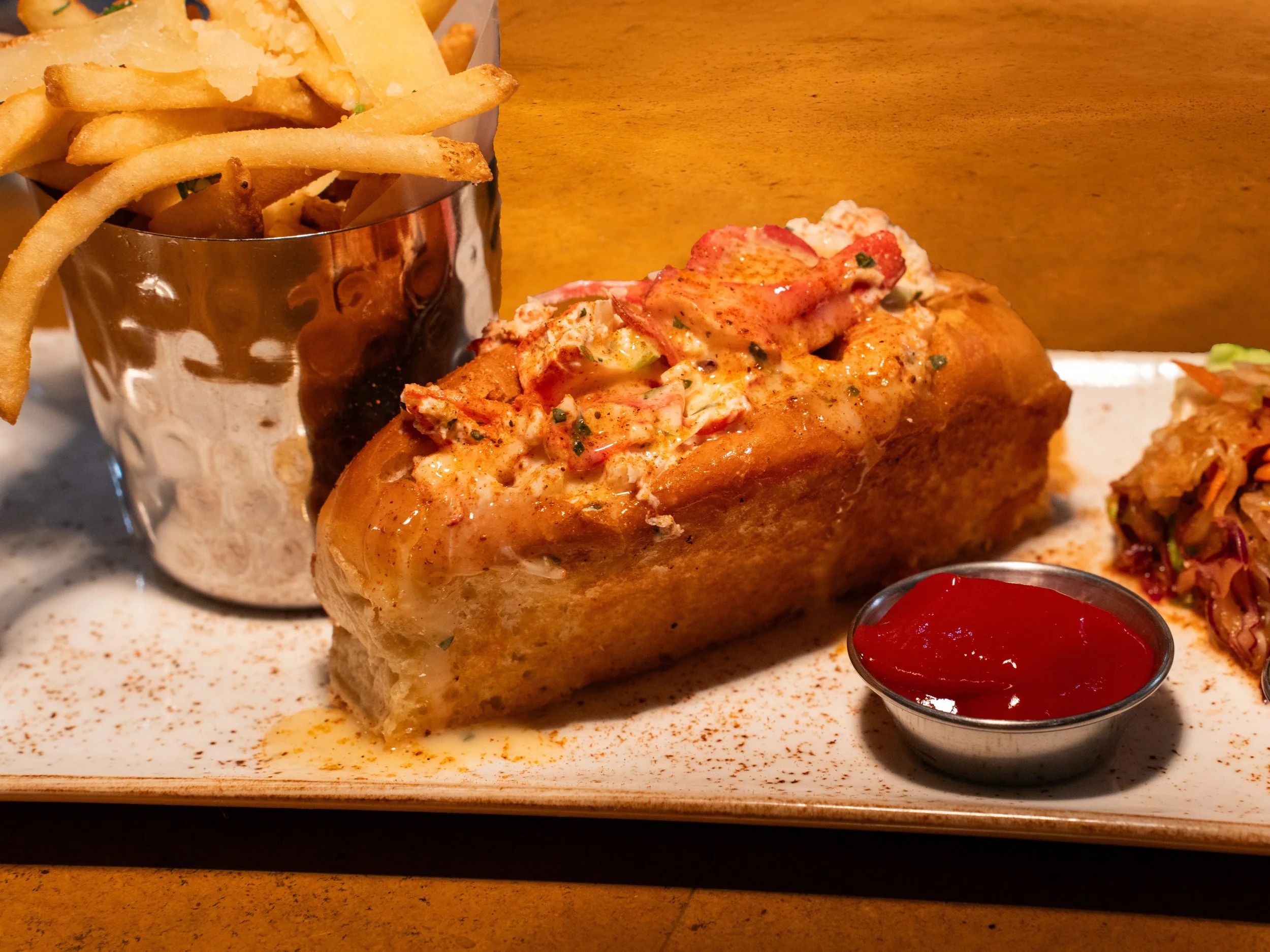A plate with a loaf of lobster roll topped with mayonnaise and seasonings, a side of French fries in a metal cup, and a small dish of ketchup, with coleslaw on the side.
