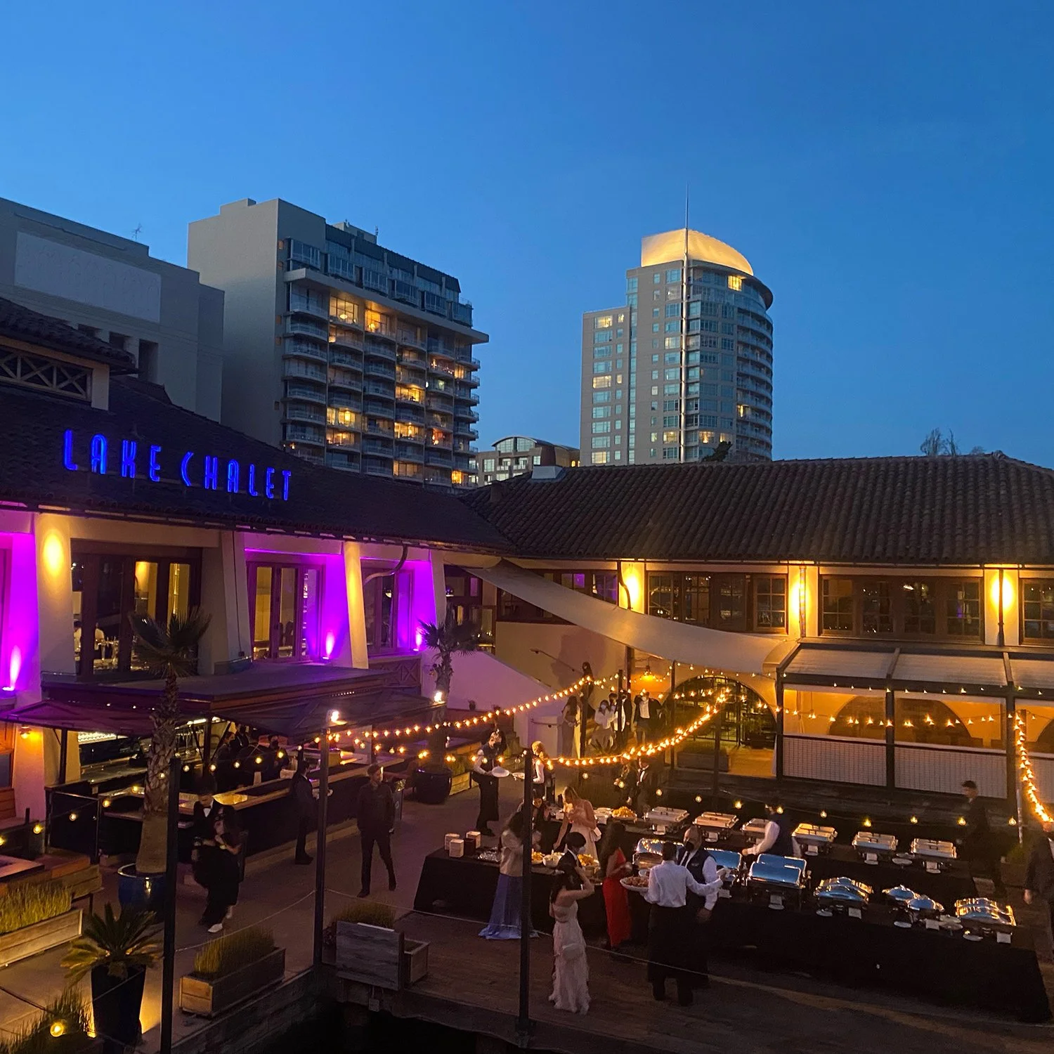 An outdoor rooftop restaurant during evening hours with string lights, people dining, and high-rise buildings in the background.