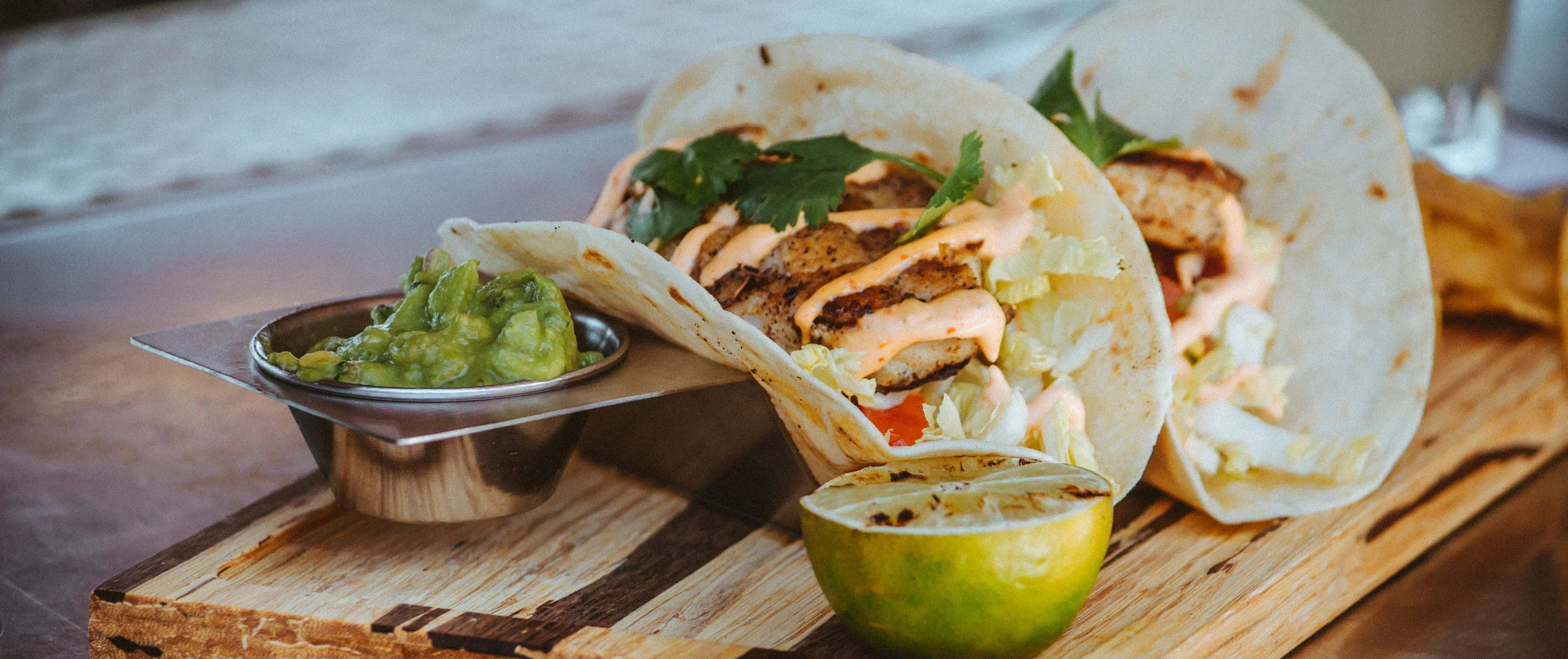 Three fish tacos with grilled fish, lettuce, and creamy sauce, served with guacamole and a grilled lime half on a wooden board.