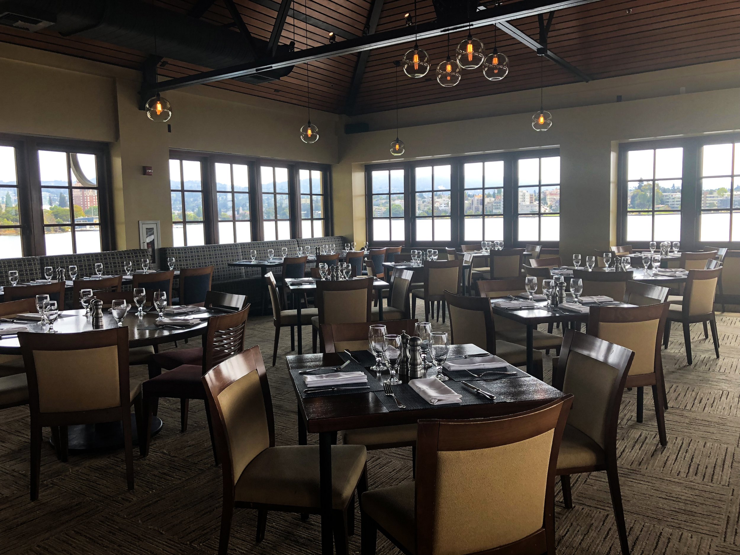 Empty restaurant with wooden tables and chairs set for dining, large windows overlooking a cityscape, and modern pendant lights hanging from a wooden ceiling.