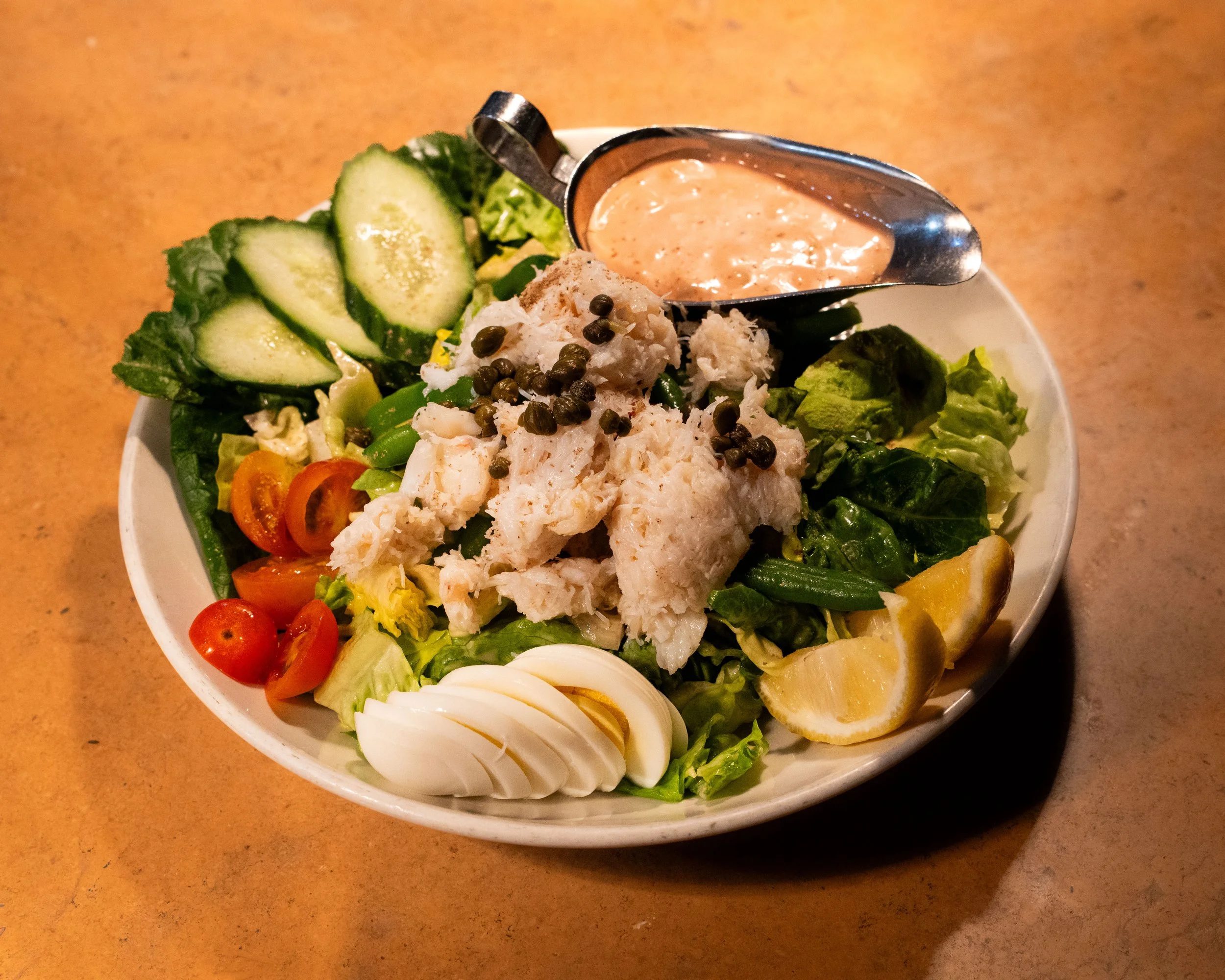 A bowl of mixed salad containing cucumber slices, cherry tomatoes, sliced hard boiled eggs, shredded chicken topped with peppercorns, green lettuce, avocado chunks, and lemon wedges, with a side of creamy dressing.