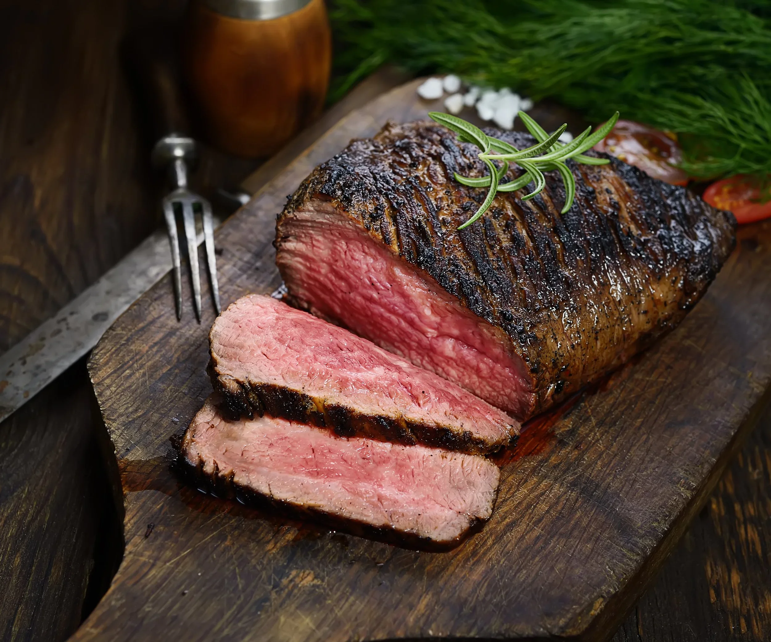 Cooked beef steak with a charred crust, sliced to show pink interior, garnished with rosemary on a wooden cutting board.