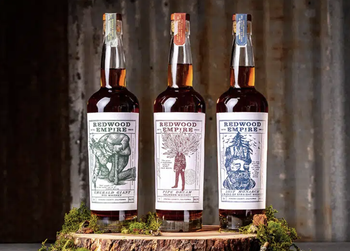 Three bottles of Redwood Empire bourbon whiskey displayed on a wooden surface with moss and bark beside them, against a wooden background.