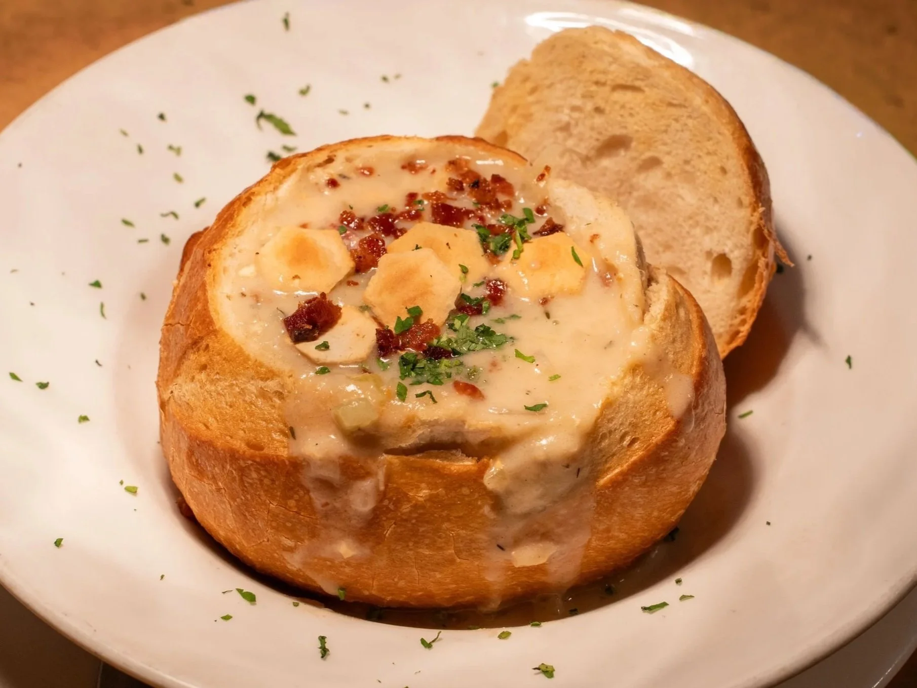Baked bread bowl filled with creamy clam chowder, garnished with chopped parsley, bacon bits, and small potato pieces, served with two slices of bread on a white plate.