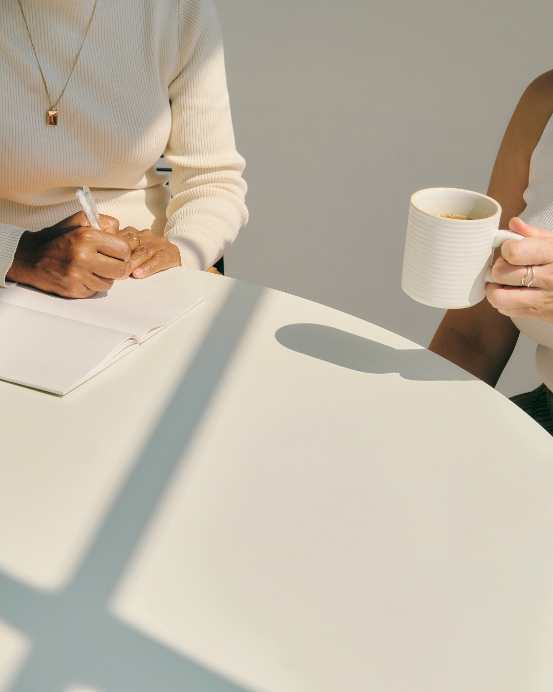 Two people sitting at a white table, one writing in a notebook, the other holding a mug of coffee.