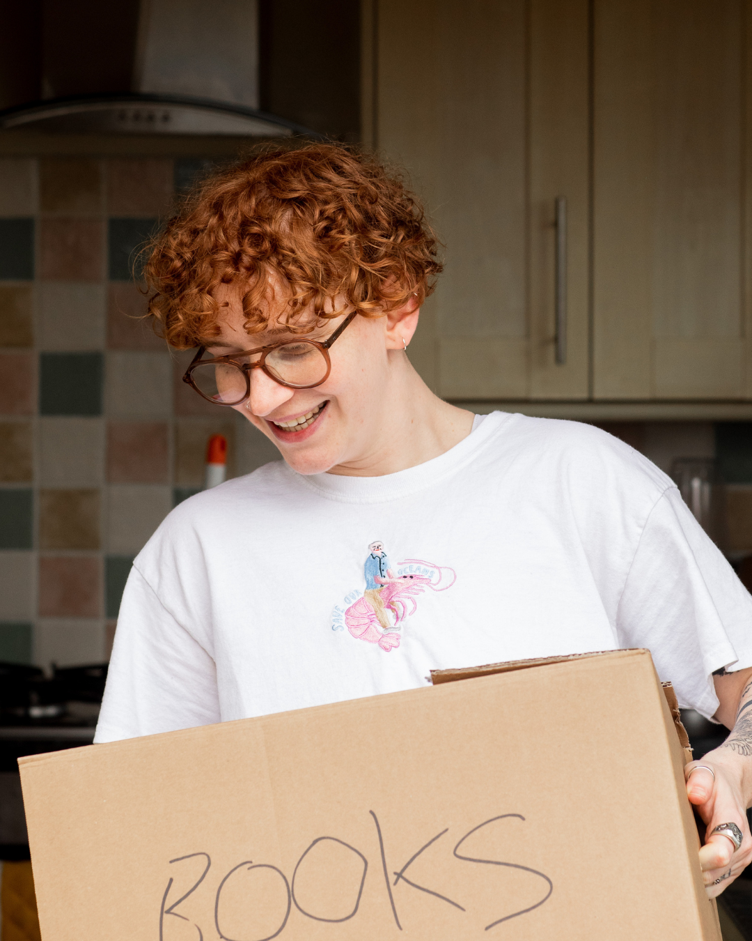 A person with curly red hair, glasses, and earrings smiling while holding a cardboard box labeled 'BOOKS' in a kitchen with tile backsplash and wooden cabinets.