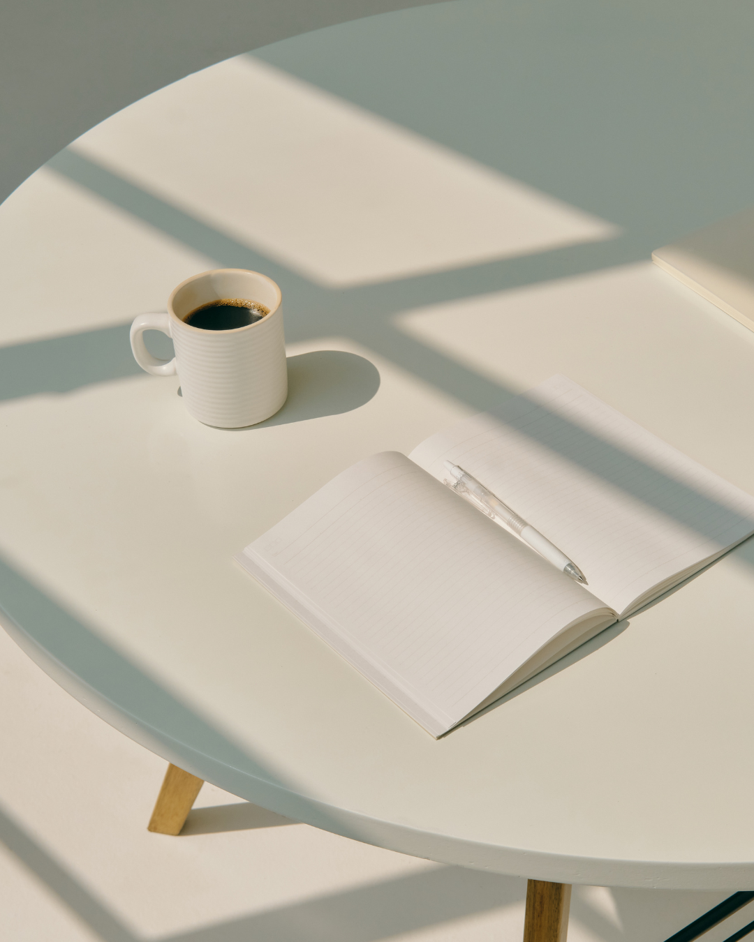 A white round table with a cup of black coffee, an open notebook with lined pages, and a clear pen placed on top, illuminated by sunlight with shadows cast across the surface.