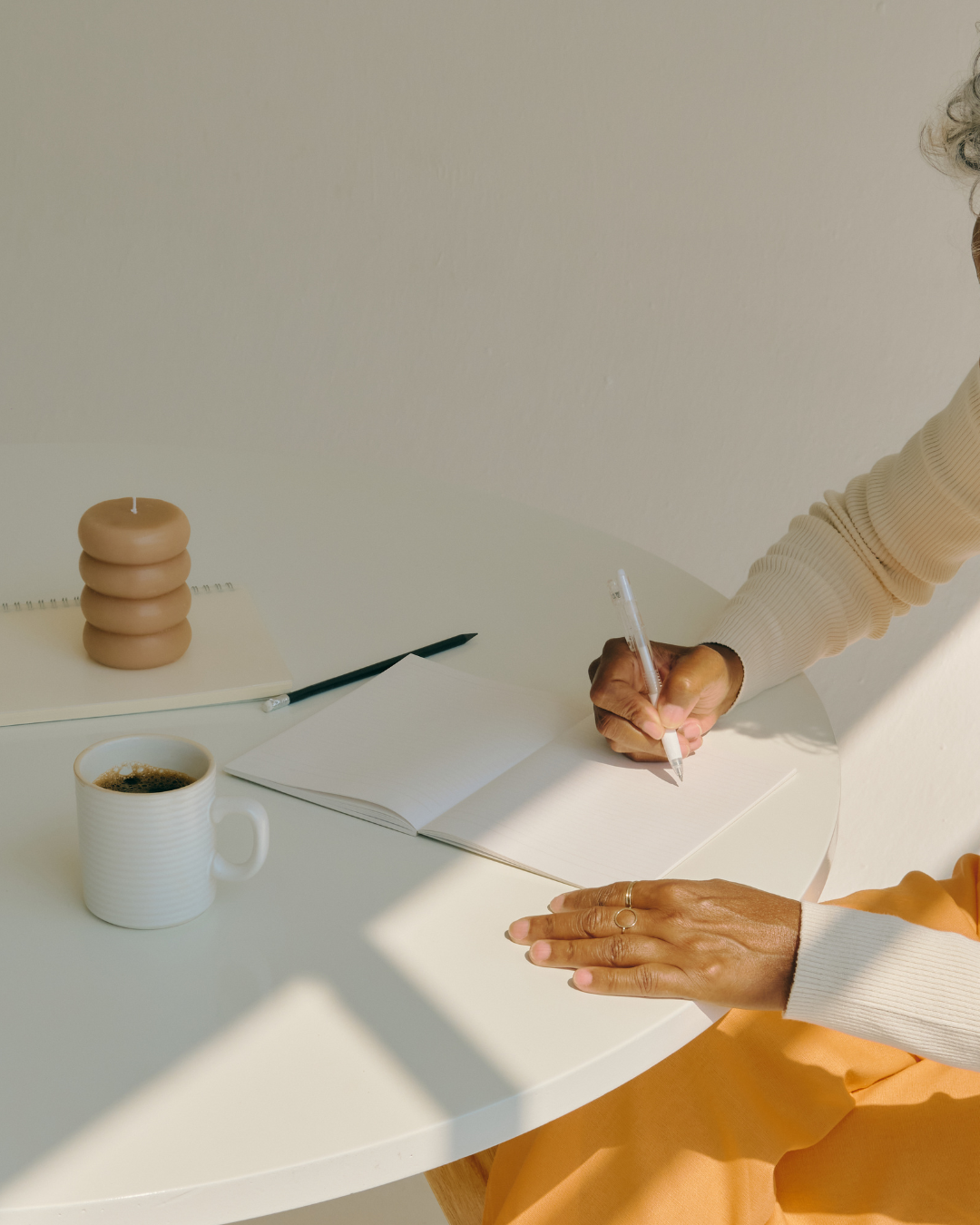 Person writing in a notebook at a white table with a cup of coffee, a black pen, a spiral notebook, and a stack of three beige candles.