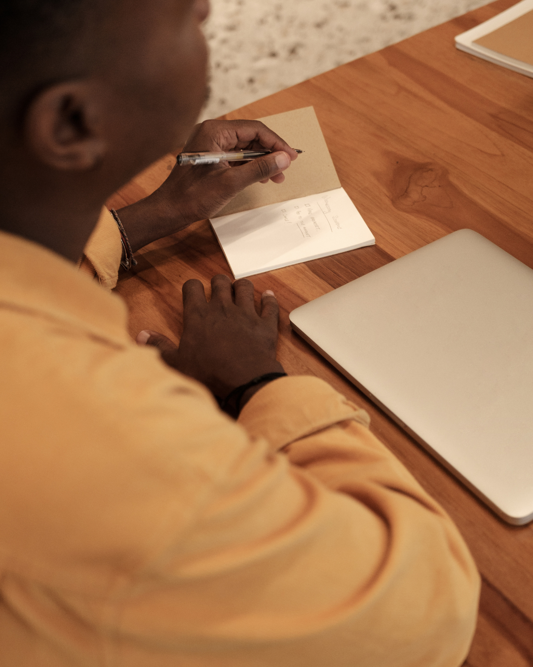 Person in a yellow sweatshirt writing in a small notebook on a wooden table, with a silver laptop and a closed notebook nearby.