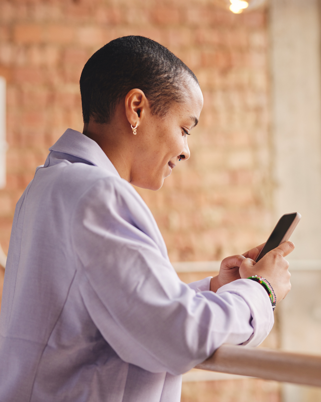 A woman with a short shaved hairstyle and earrings, wearing a light purple shirt, smiling and looking at her phone inside a room with a brick wall background.