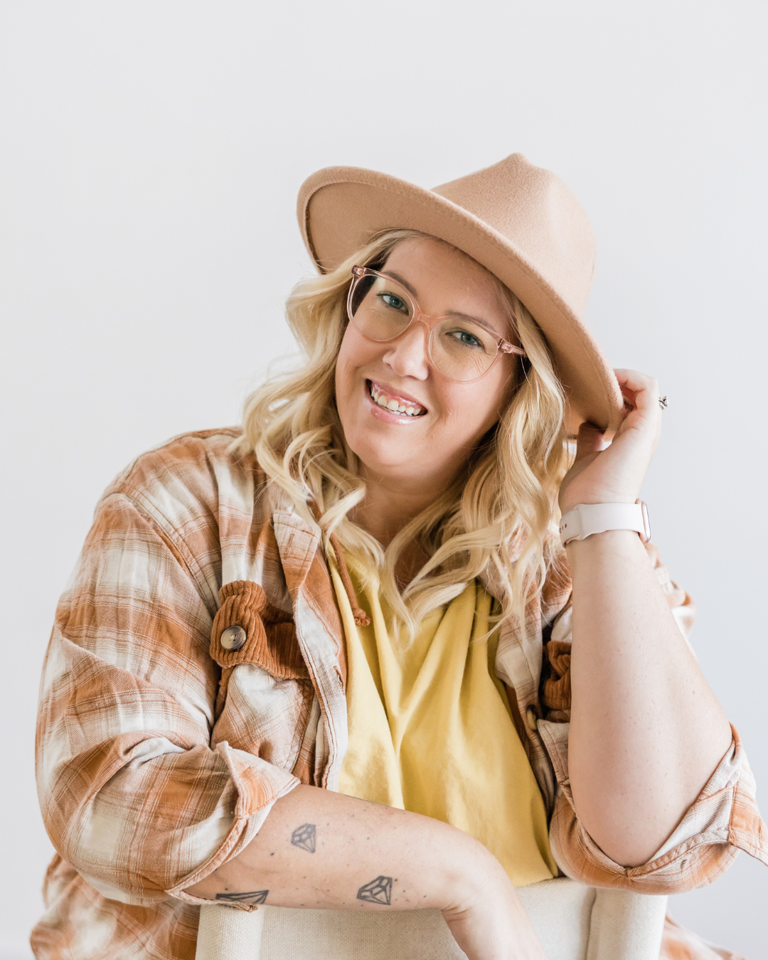 A smiling woman with blonde hair, wearing a beige wide-brim hat, glasses, a yellow shirt, and a plaid jacket, sitting against a white background.