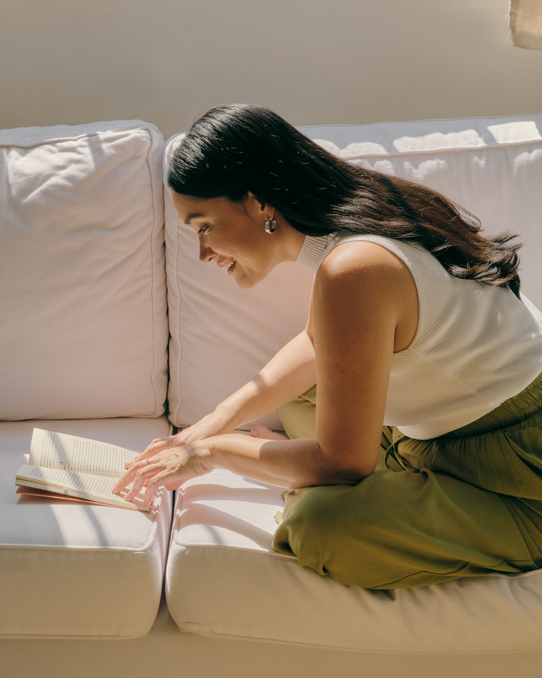 A woman with long black hair, wearing a sleeveless white top and olive-green pants, is sitting on a white sofa, reading a book, smiling, with sunlight streaming in.