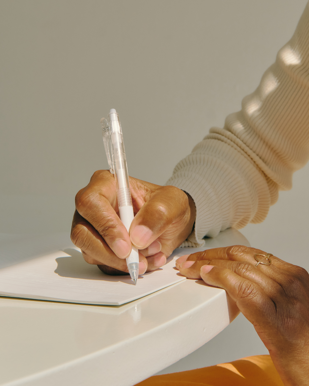 A person writing on a notepad with a pen, wearing a ring on their left hand, and a beige ribbed long-sleeve shirt.
