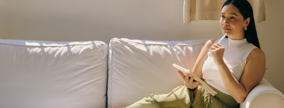 Woman sitting on a couch with a notebook, holding a pen, in a well-lit room with a beige curtain