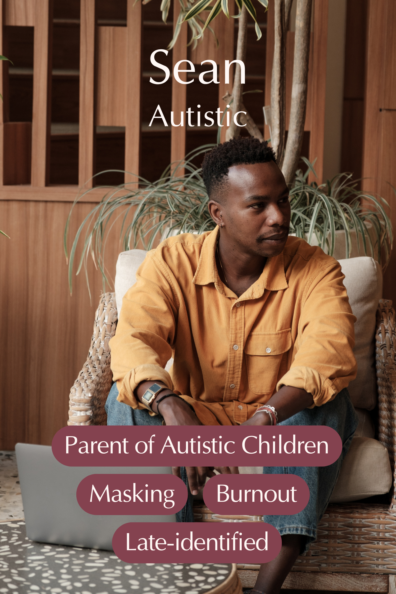 A young man with dark skin and short curly hair, wearing a mustard-colored shirt, sitting on a wicker chair with a tablet on a table in front of him. Behind him are potted plants and wooden shelving.