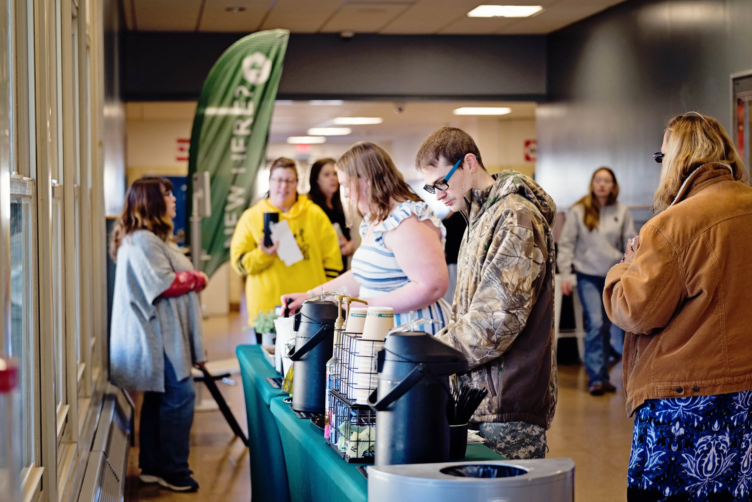 People in line at a coffee or beverage stand inside a building with a green banner.