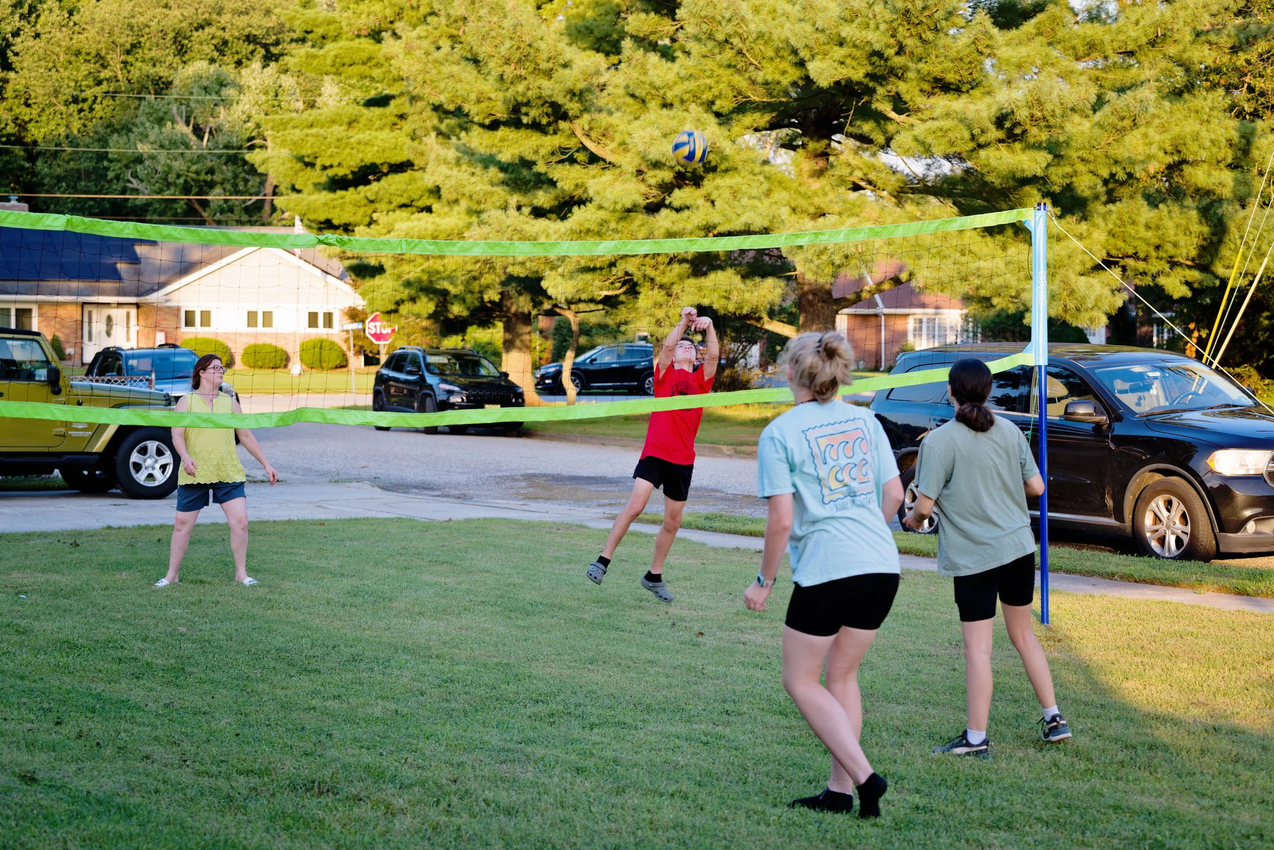 People playing volleyball outdoors in a grassy yard with trees and parked cars in the background.