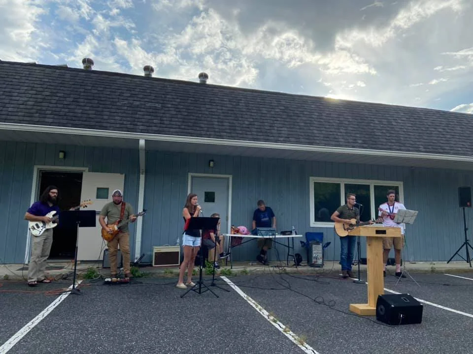 A group of seven musicians performing outdoors near a blue building with a parking lot in the foreground.