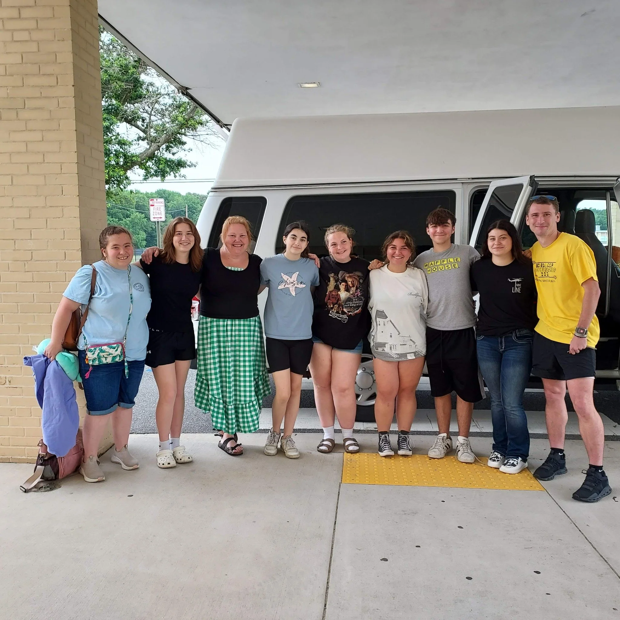 A group of nine people standing in front of a white van under a covered area, smiling and posing for the photo. Some are wearing casual clothing, including t-shirts, shorts, and skirts.