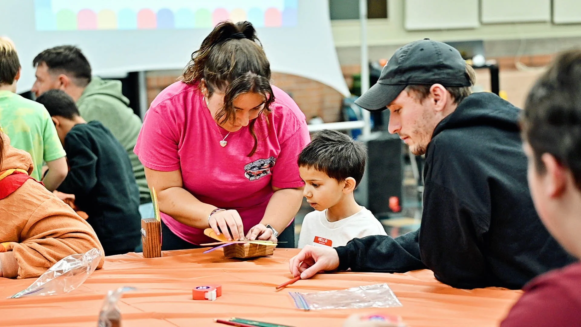 Adults and children working together on a craft project at a table covered with an orange tablecloth, in a community center or classroom setting.