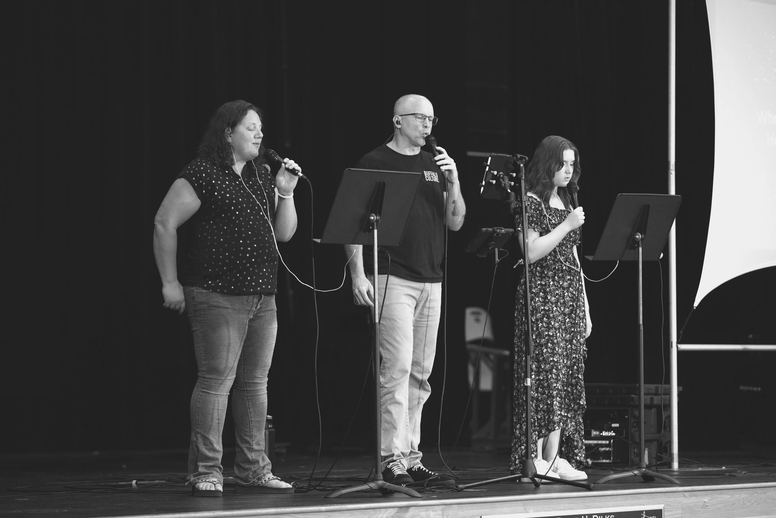 Three people singing or speaking into microphones on stage at a presentation or performance.