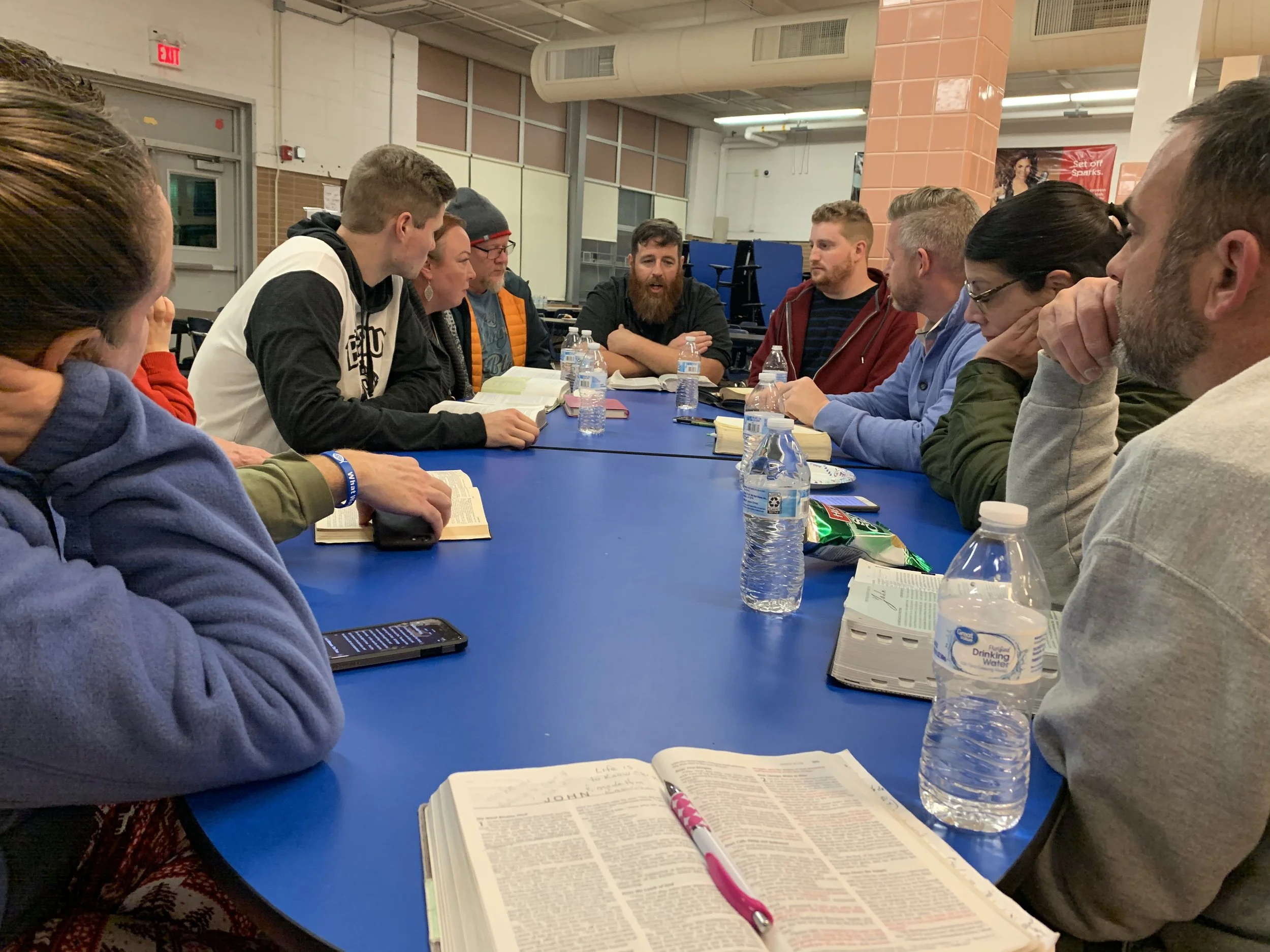 People sitting around a blue table engaged in discussion with books, water bottles, and snacks on the table.