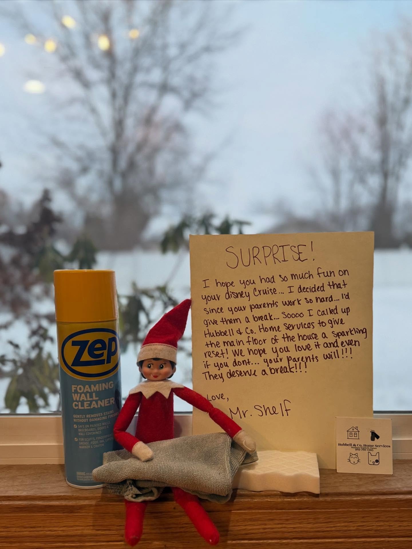 A doll elf wearing a red outfit and Santa hat, sitting with folded cloth and a can of Zep foam cleaner next to a handwritten note on a windowsill. Outside the window, there are trees and fog.