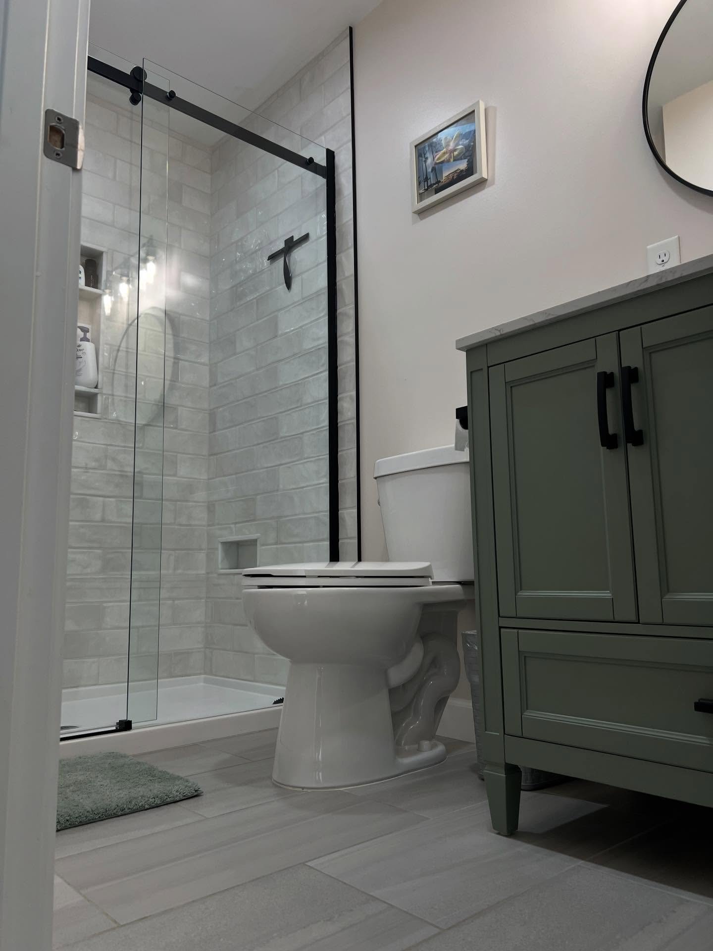 Bathroom with a walk-in shower with glass doors, a white toilet, and a green bathroom vanity with black handles. There is a small framed picture and a mirror on the wall.