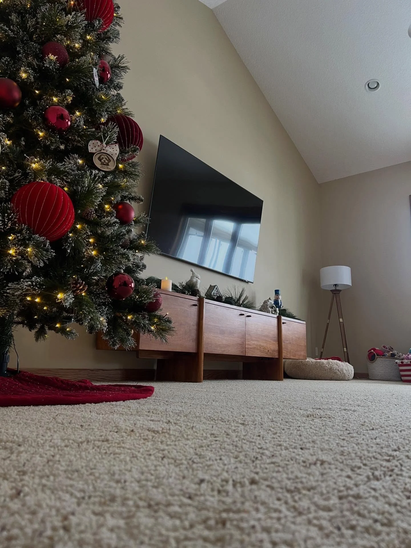 A decorated Christmas tree with red ornaments and white lights in a cozy living room. A wooden console beneath the TV holds holiday decorations. To the right, there's a standing lamp, a pet bed, and a basket filled with toys.