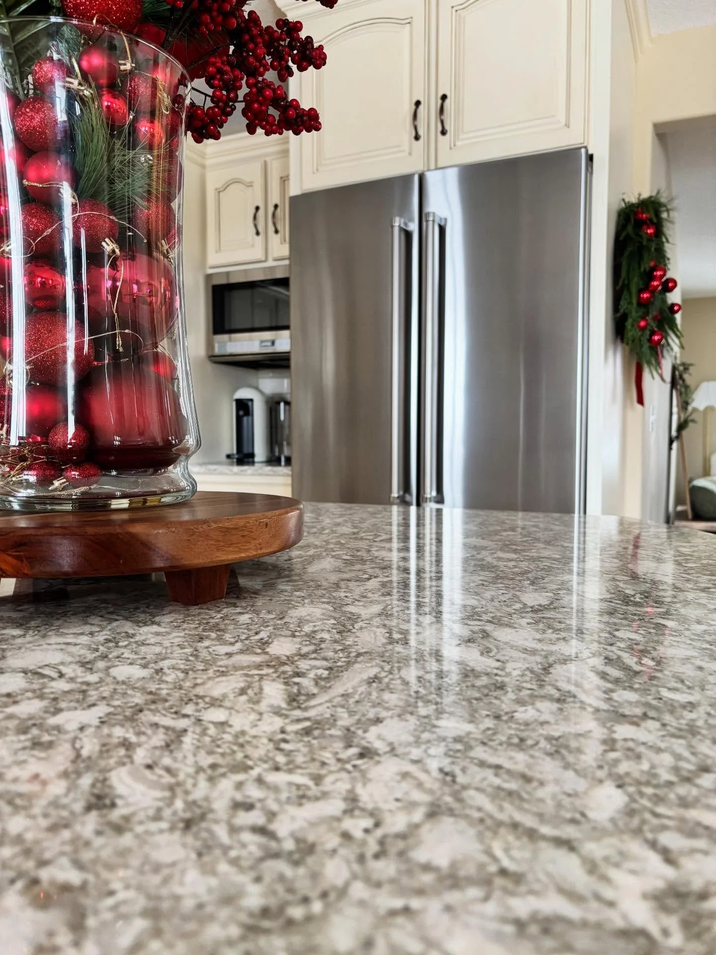 Close-up of a granite kitchen countertop with a clear glass vase filled with red holiday ornaments and greenery, placed on a wooden tray. In the background, there are white kitchen cabinets, a stainless steel refrigerator, and holiday decorations on 