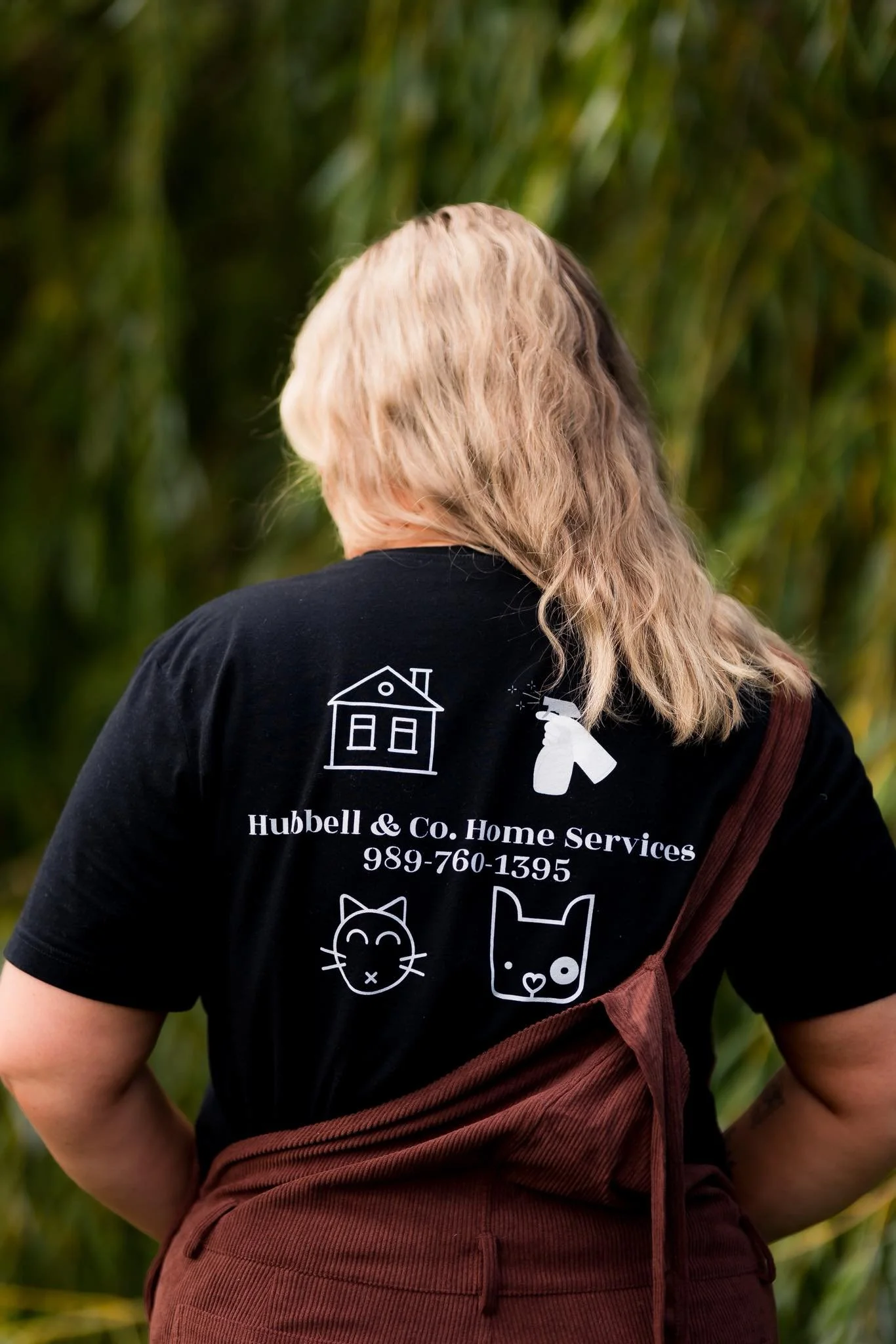 Back of a woman wearing a black t-shirt with white graphics and text advertising Hubbell & Co. Home Services, standing outdoors with a green plant background.