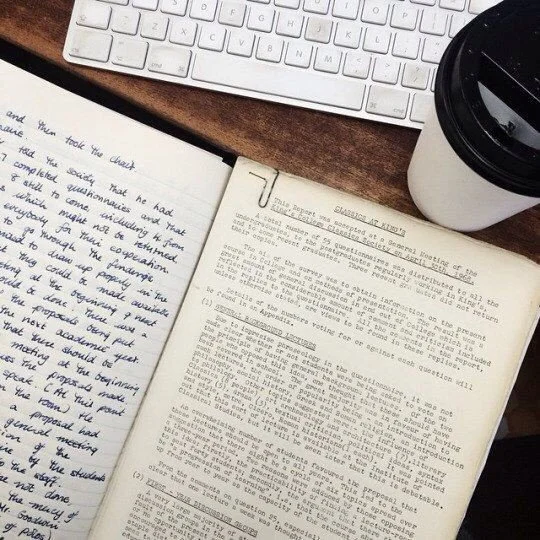 Open notebook with handwritten notes, a white keyboard, a black coffee cup, and a brown desk surface.
