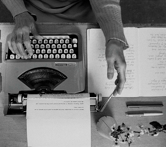 A person using a typewriter and a computer keyboard on a wooden desk, with papers, pens, and a small potted plant.