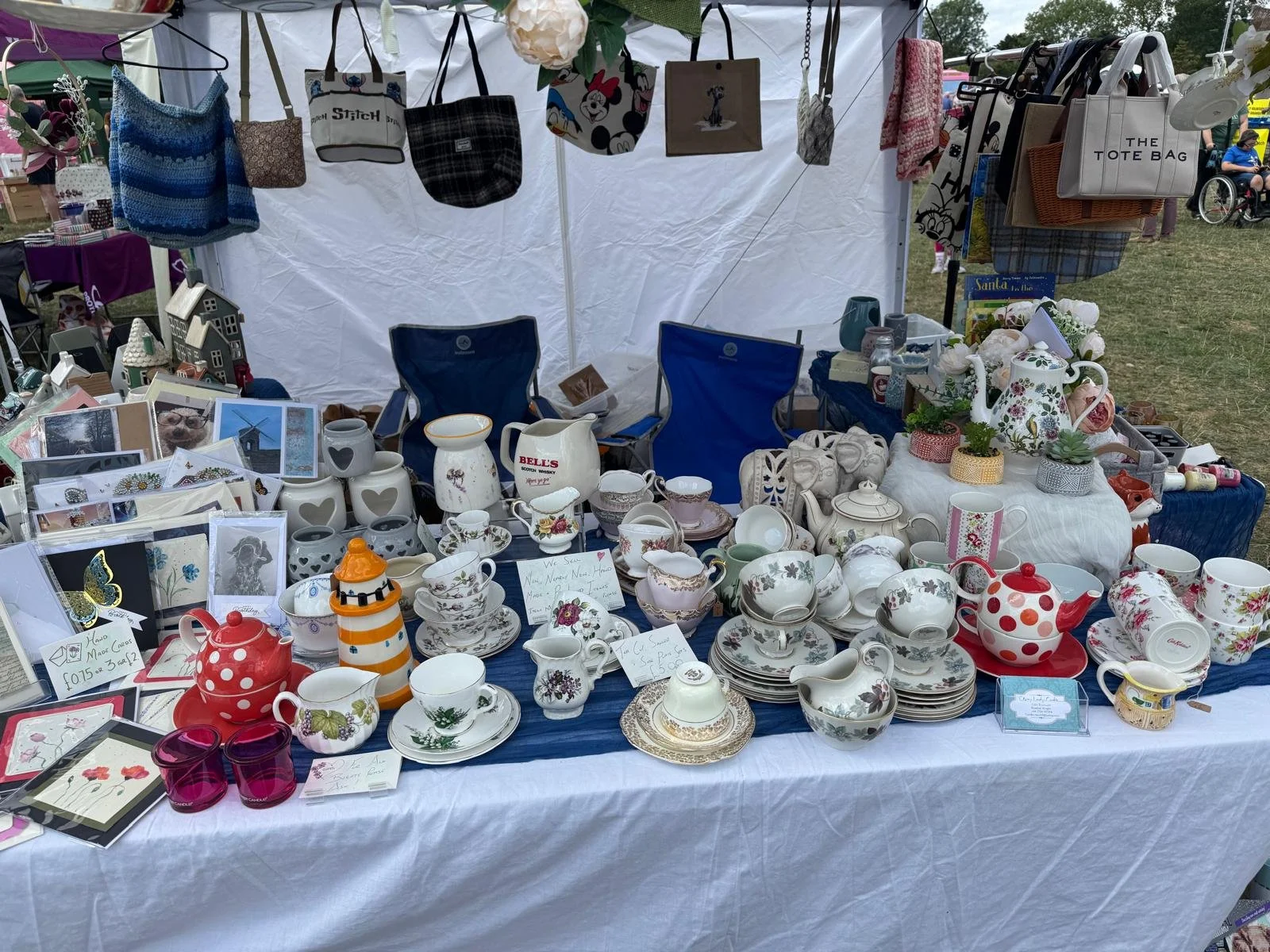 A table at an outdoor flea market display with various ceramics, teapots, cups, and decorative items. Behind the table, hanging bags and tote bags are displayed under a white canopy with people and tents visible in the background.