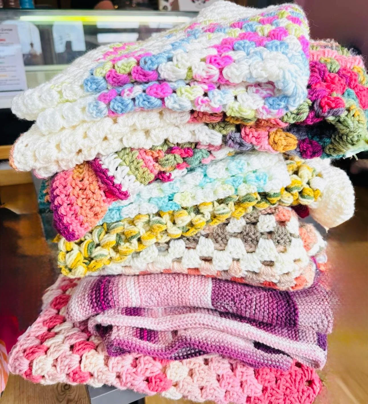 Stacked colorful crocheted and knitted blankets in pink, white, yellow, green, blue, purple, and beige placed on a table.