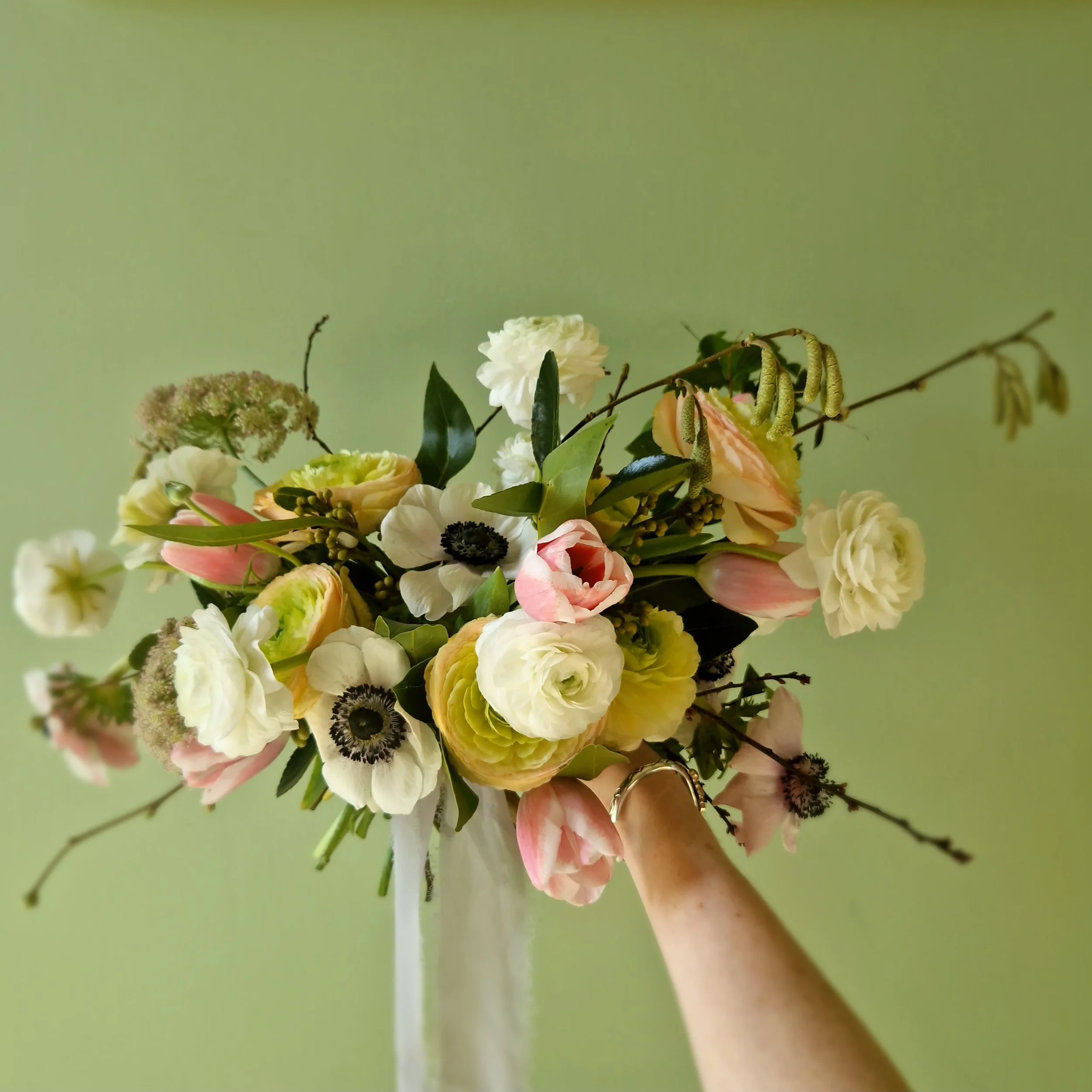 Hand holding a bouquet of various flowers, including white, pink, and yellow blooms, against a pale green background.