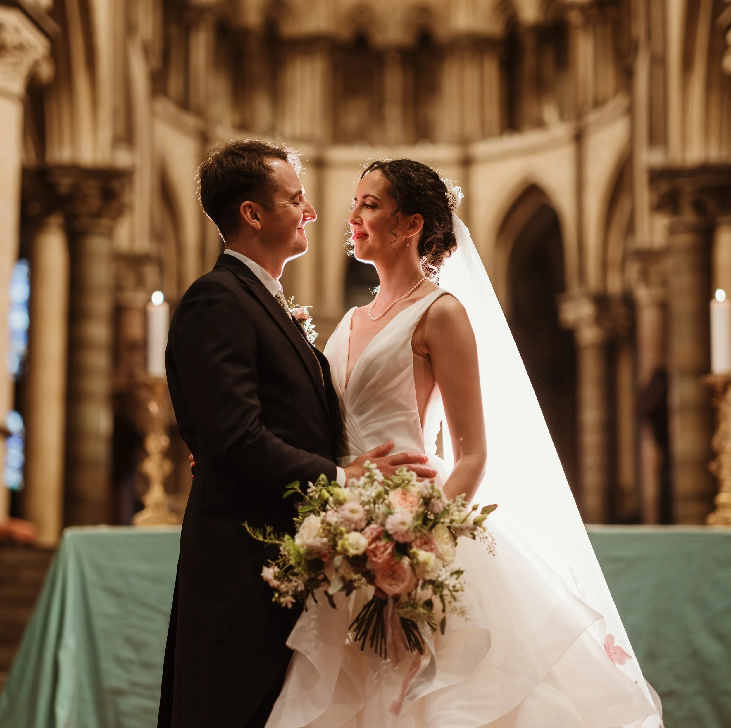 A bride and groom standing together at the alter of Canterbury Cathedral with the bride holding a bouquet of seasonal pink and white flowers.