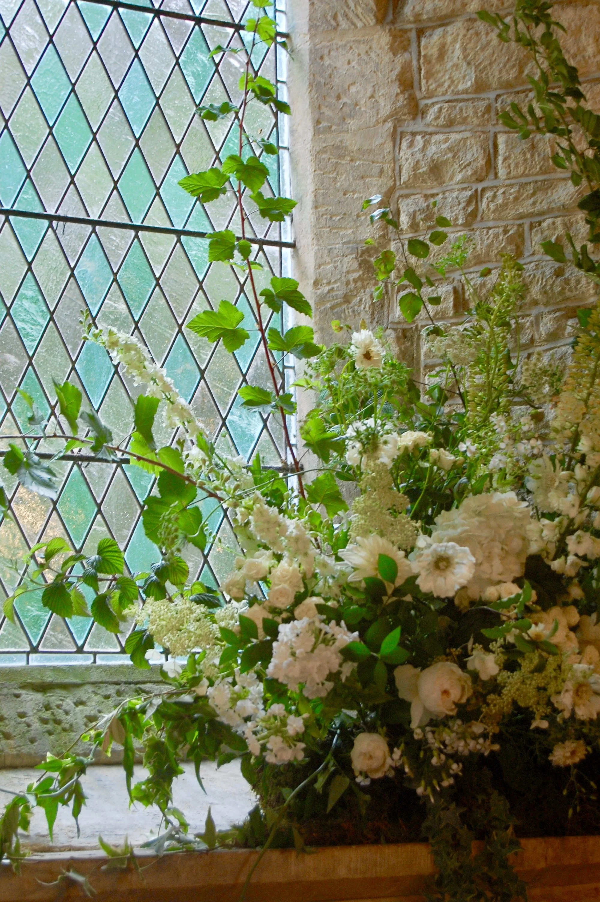 A large arrangement of white flowers, including roses and other varieties, placed on a windowsill with a diamond-patterned glass window and a brick wall in the background.