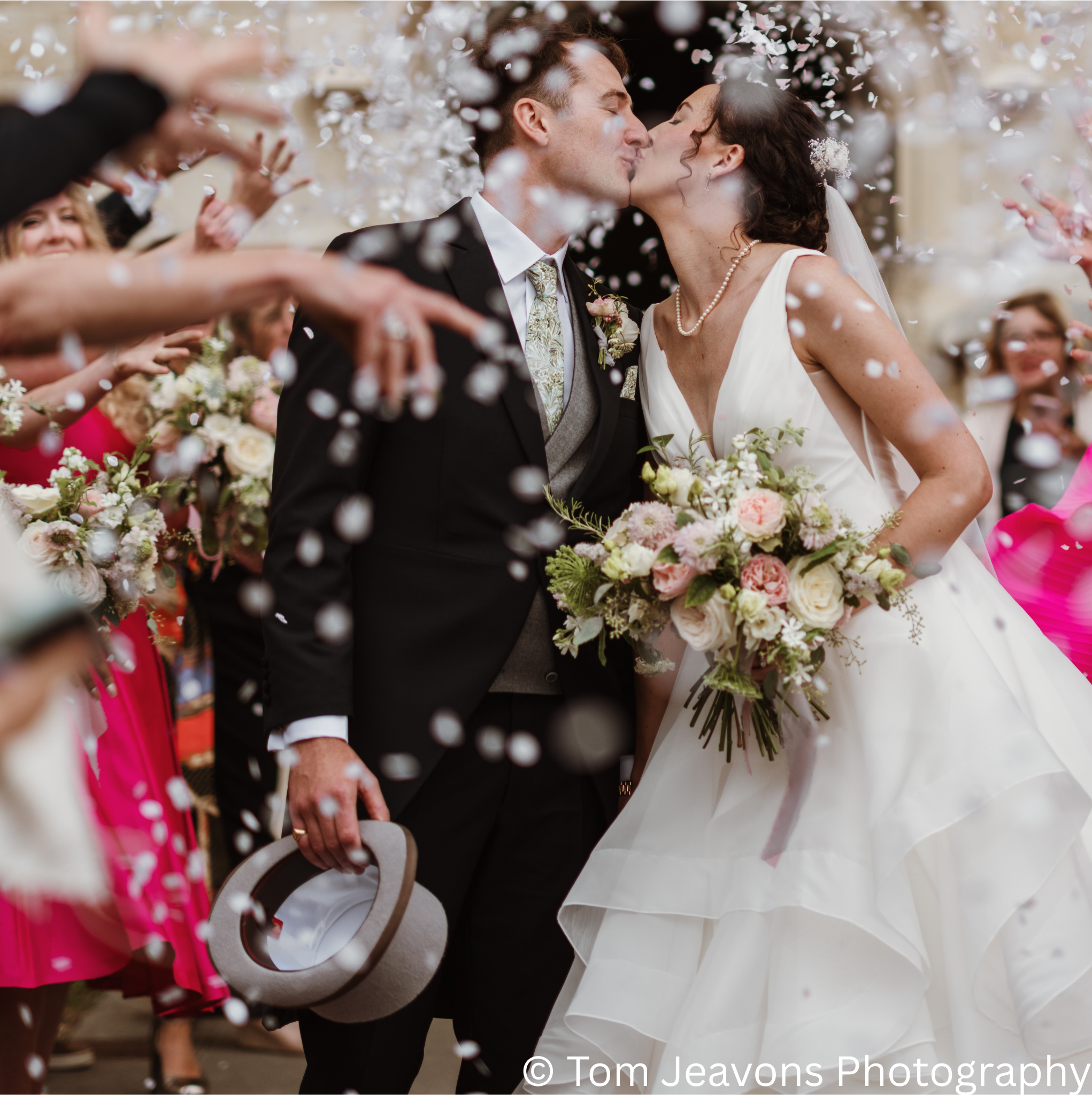 A bride and groom share a kiss during their wedding celebration, surrounded by guests throwing confetti and holding flowers, with the groom holding a hat.