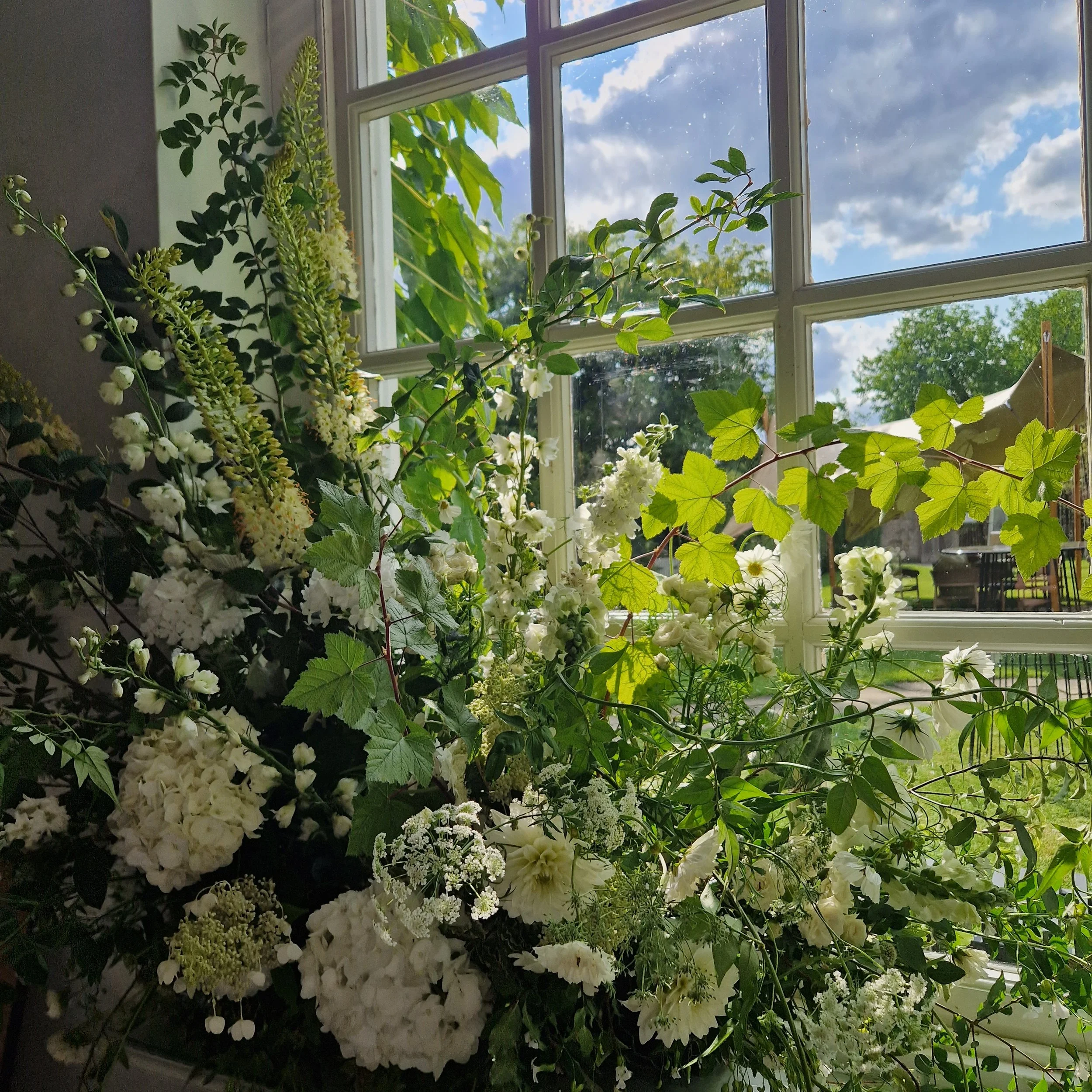 A bouquet of white flowers and greenery on a windowsill with a view of a garden outside, blue sky, and clouds.
