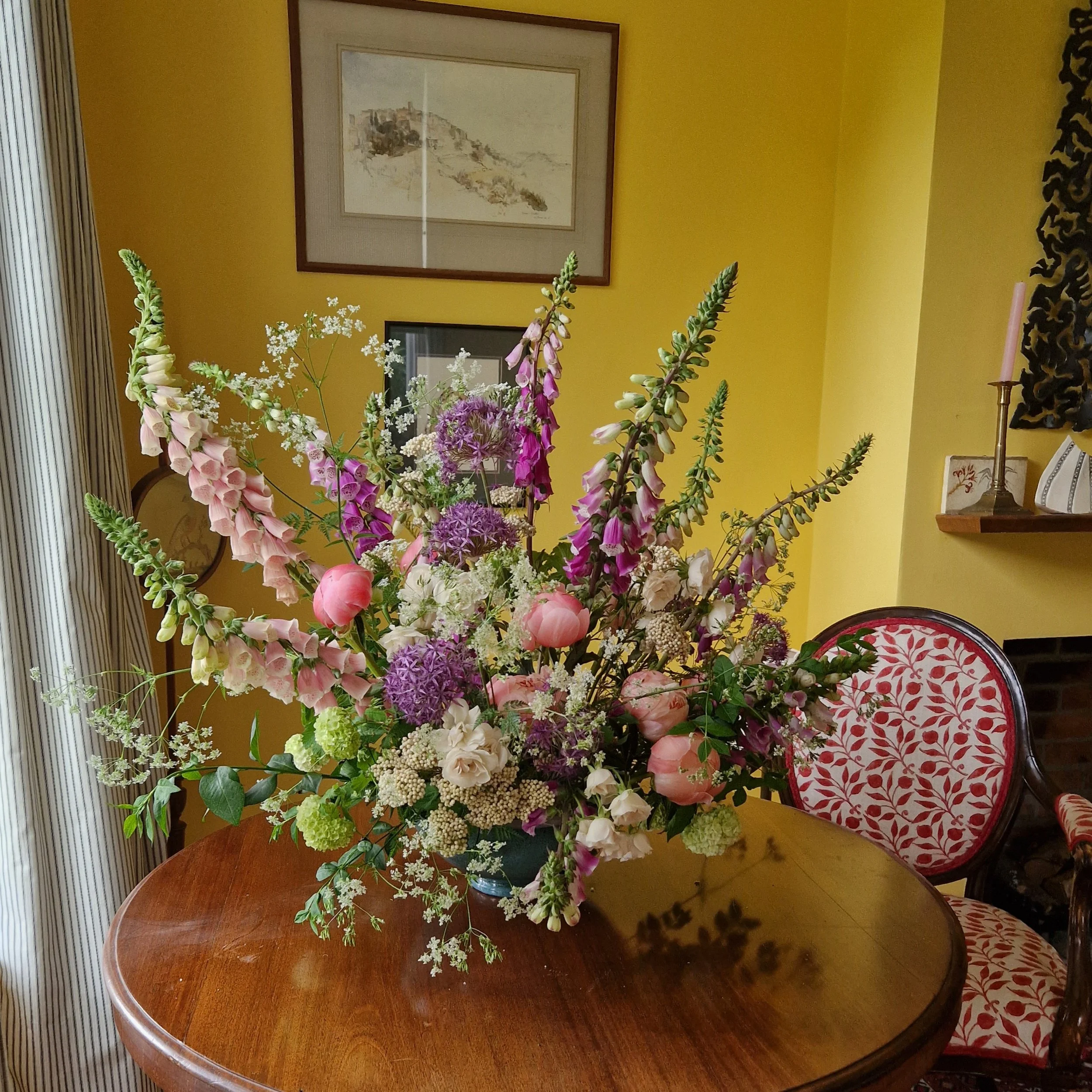 A large bouquet of pink, purple, white, and green flowers arranged on a wooden table in a cozy room with yellow walls, a framed picture, a decorative wall hanging, and a patterned chair.