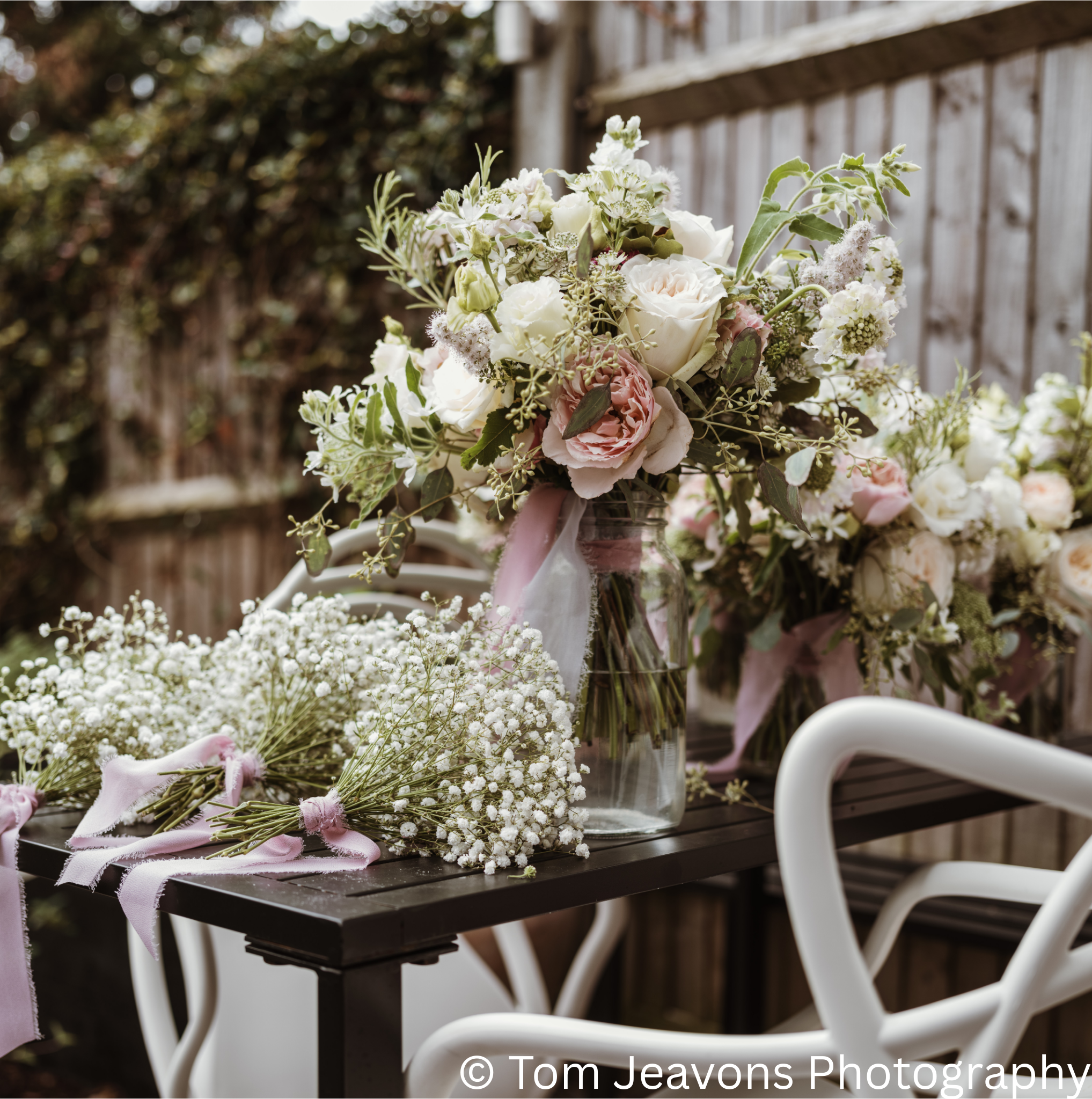 Vase with a floral arrangement of white and pink roses, green foliage, and small white flowers, on a black table outdoors.