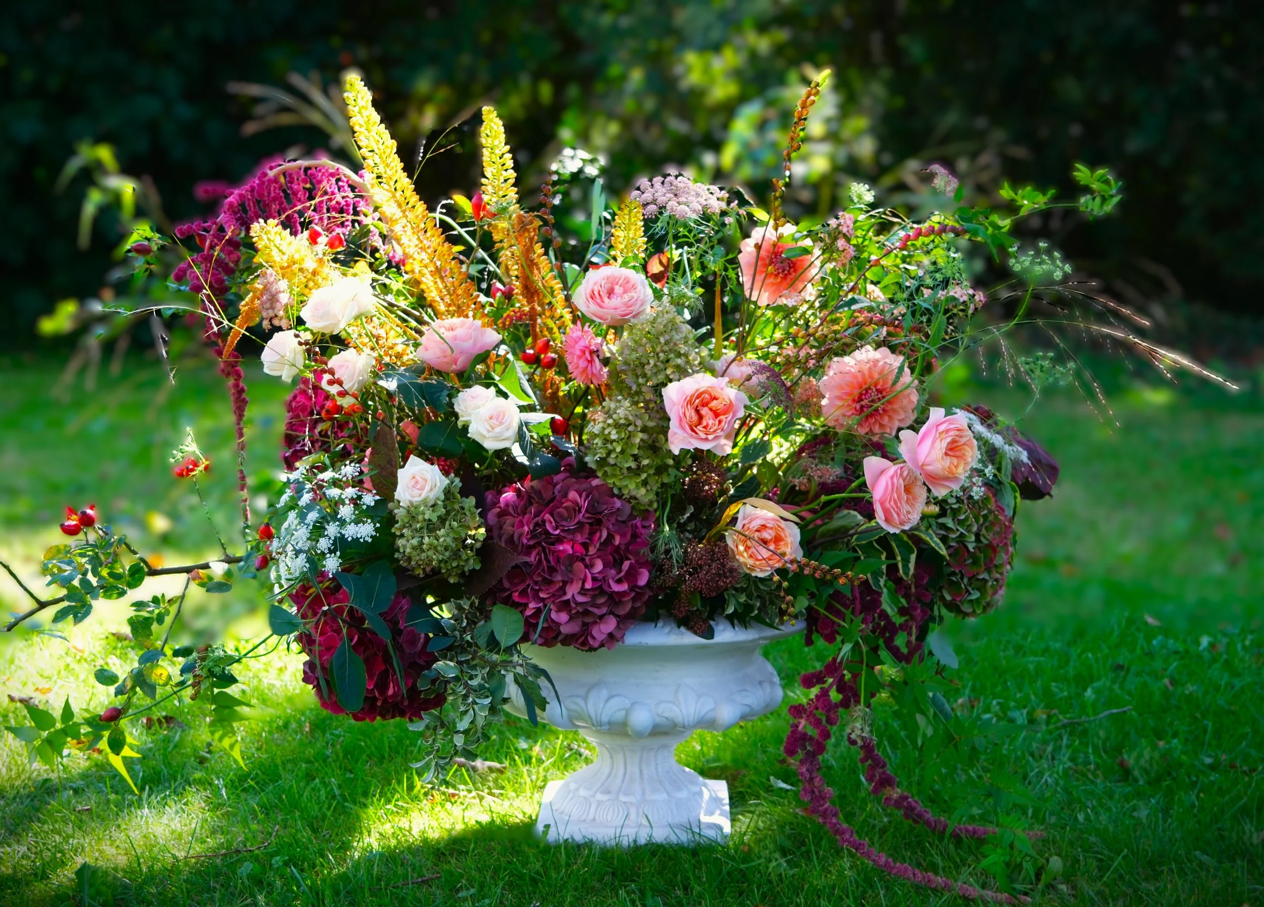 Colourful autumnal flower arrangement in a white urn in a garden setting.  Warm, dappled sunlight highlights the florals.