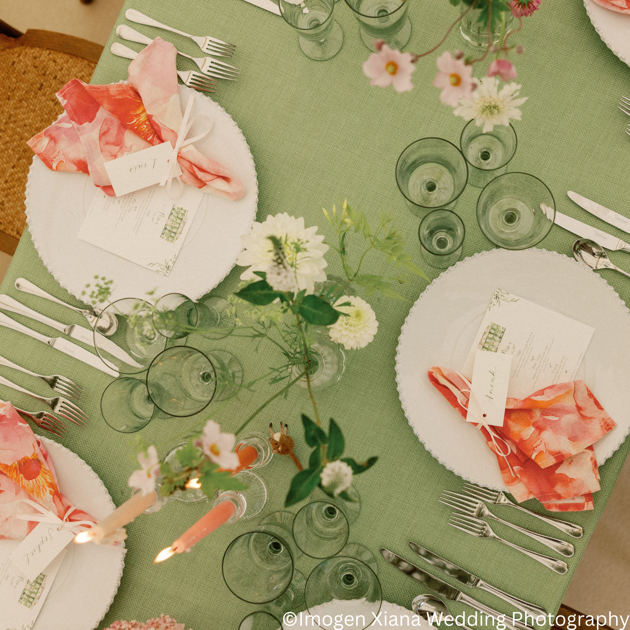 Table setting with white plates, pink floral napkins, silverware, multiple glasses, and floral centerpieces with pink and white flowers on a green tablecloth.