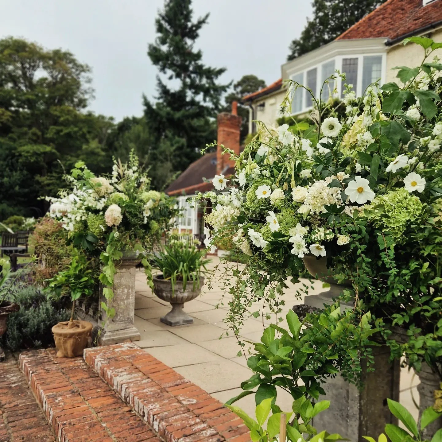 View of a lush, flowering garden with white flowers in urns and pots in front of a large, traditional house with a red-tiled roof and bay windows, surrounded by mature trees.