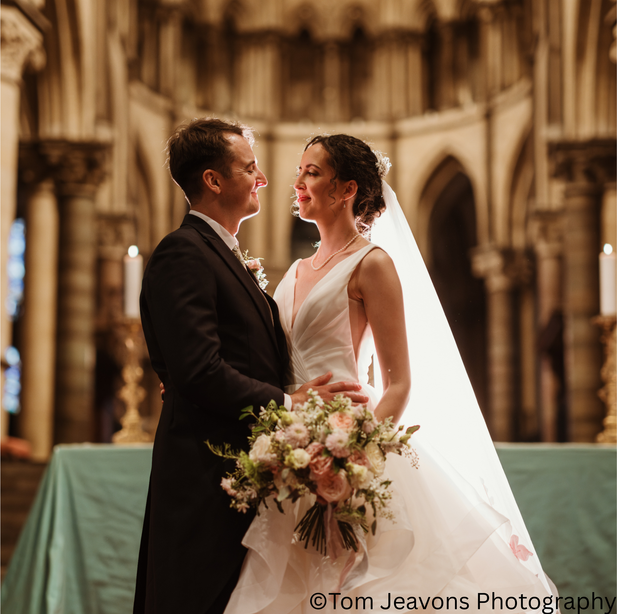 A bride and groom facing each other and smiling inside a church, with the bride holding a bouquet of flowers and wearing a white wedding gown with a veil, and the groom in a black tuxedo.