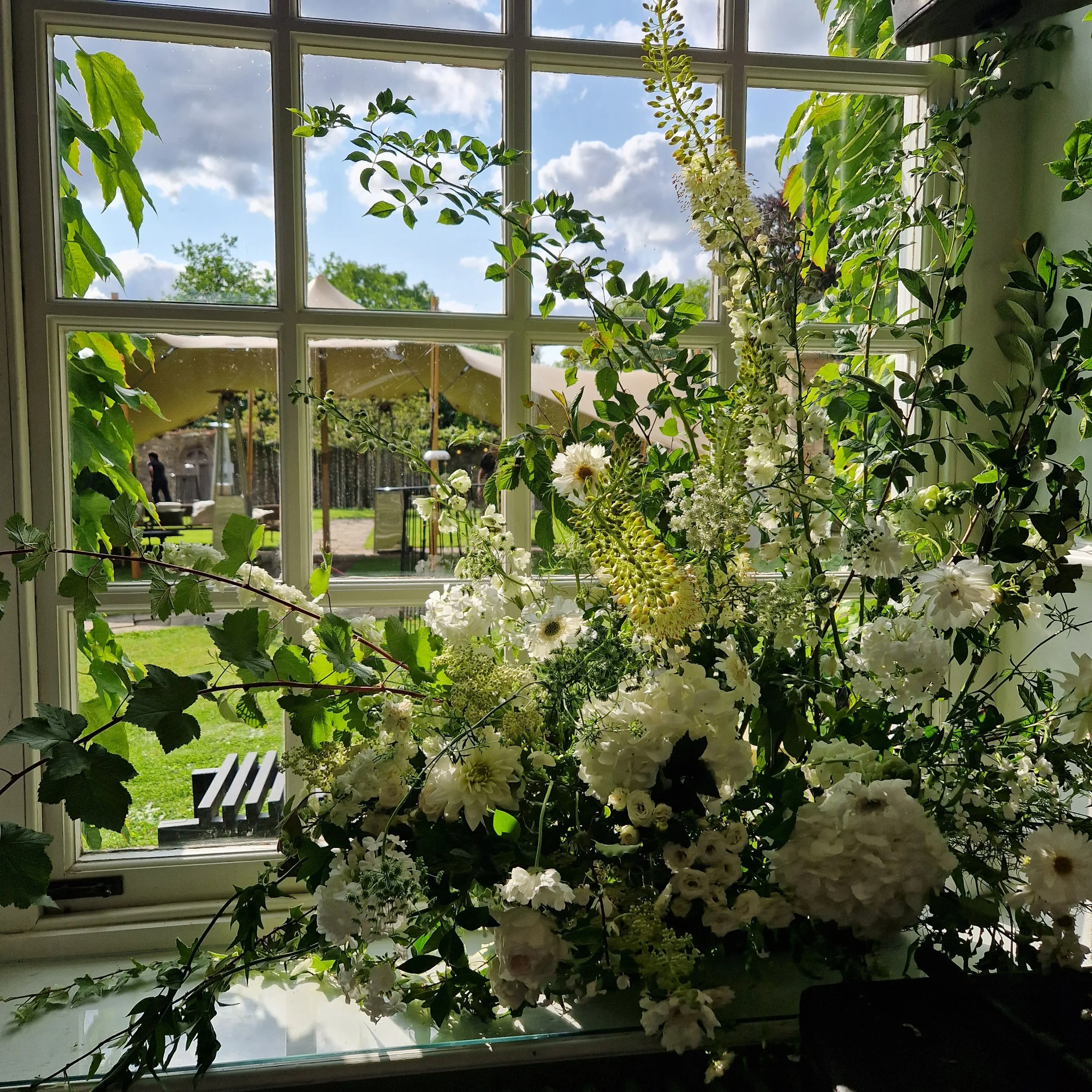 A large bouquet of white flowers and green foliage placed on a windowsill inside a house, with a view of a sunny garden and blue sky outside.